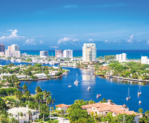 Aerial view of Fort Lauderdale, featuring a winding blue waterway lined with boats and bordered by lush greenery, urban buildings, and a bright, clear sky.