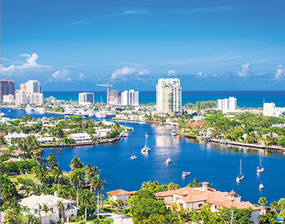 Aerial view of Fort Lauderdale, featuring a winding blue waterway lined with boats and bordered by lush greenery, urban buildings, and a bright, clear sky.