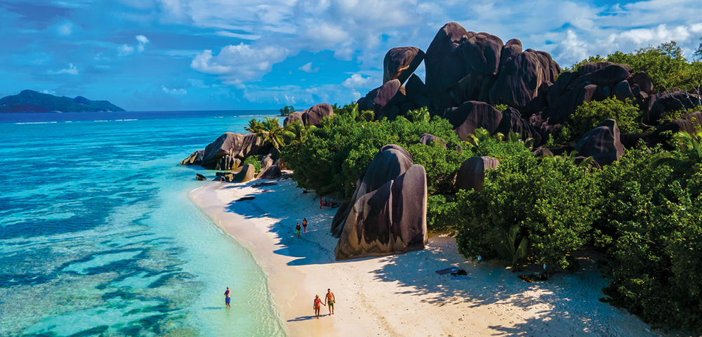 A tropical beach with clear turquoise water and white sand, lined with lush green foliage and large, unique rock formations. People stroll along, evoking tranquility.