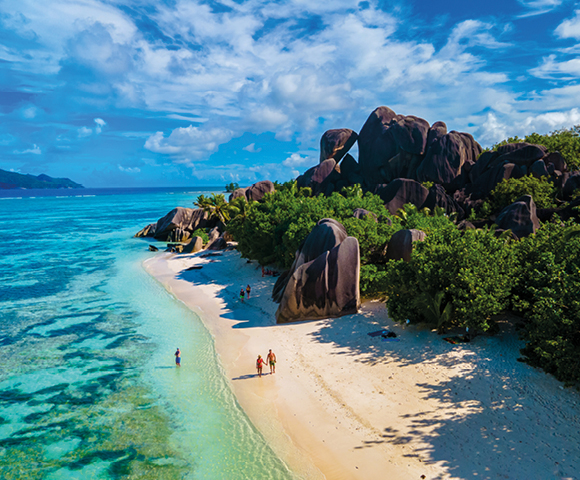 A tropical beach with clear turquoise water and white sand, lined with lush green foliage and large, unique rock formations. People stroll along, evoking tranquility.