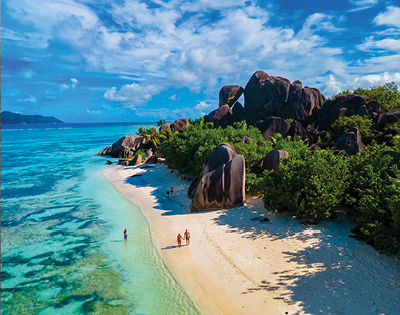 A tropical beach with clear turquoise water and white sand, lined with lush green foliage and large, unique rock formations. People stroll along, evoking tranquility.