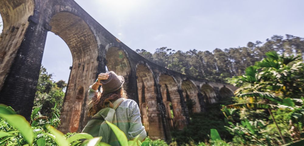 Woman wearing a hat and backpack looks up at the Nine Arches Bridge in a lush, green forest. The sun shines brightly, creating a serene atmosphere.