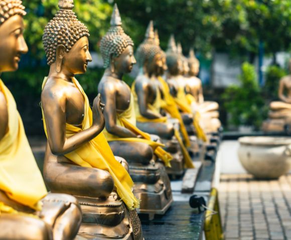 A row of serene bronze Buddha statues adorned in yellow robes sit in a tranquil garden setting, conveying peace and harmony against a backdrop of lush greenery.