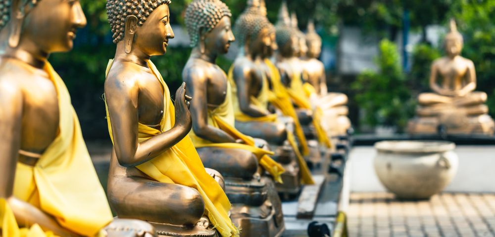 A row of serene bronze Buddha statues adorned in yellow robes sit in a tranquil garden setting, conveying peace and harmony against a backdrop of lush greenery.