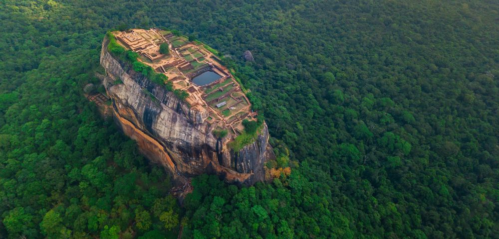 Aerial view of Sigiriya, a historical fortress in Sri Lanka, perched atop a massive rock surrounded by lush green forests under a clear sky.