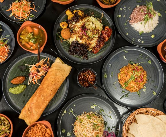 A variety of colorful Indian dishes on black plates, including dosa, rice, vegetables, and curries, creating a vibrant and appetizing spread.