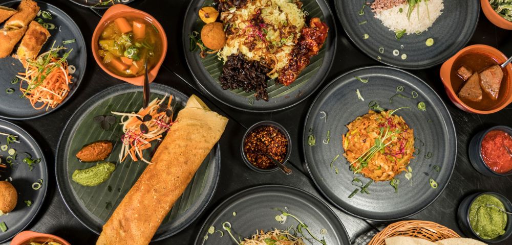 A variety of colorful Indian dishes on black plates, including dosa, rice, vegetables, and curries, creating a vibrant and appetizing spread.