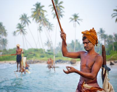 A man in traditional attire fishes on a stilt over the ocean near a tropical shoreline. Others fish in the background. Tall palm trees sway nearby.