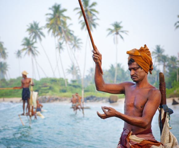 A man in traditional attire fishes on a stilt over the ocean near a tropical shoreline. Others fish in the background. Tall palm trees sway nearby.