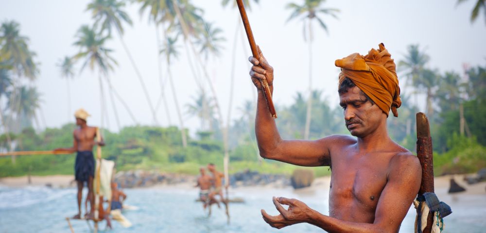 A man in traditional attire fishes on a stilt over the ocean near a tropical shoreline. Others fish in the background. Tall palm trees sway nearby.