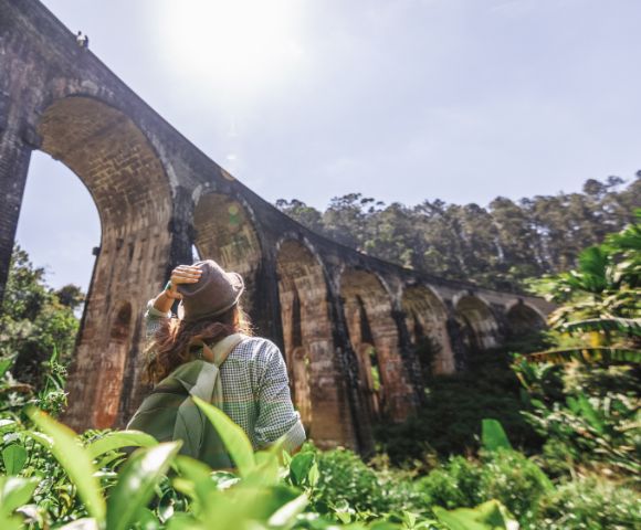 Woman wearing a hat and backpack looks up at the Nine Arches Bridge in a lush, green forest. The sun shines brightly, creating a serene atmosphere.