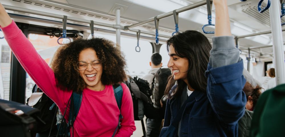 Two women, smiling and laughing, stand on a bus holding onto handrails. One wears a bright pink sweater; the mood is joyful and lively.