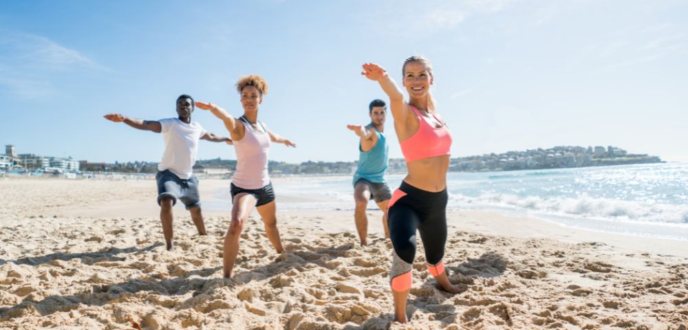 A lively group of four people exercises in the sand on a sunny beach. They stretch with arms extended, ocean waves and a clear sky in the background.