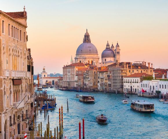 Venice's Grand Canal at sunset, with boats and gondolas in the water, surrounded by historic buildings. The sky is pink and orange, creating a serene, romantic atmosphere.