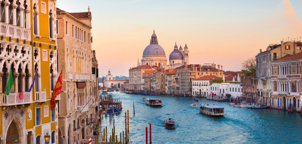 Venice's Grand Canal at sunset, with boats and gondolas in the water, surrounded by historic buildings. The sky is pink and orange, creating a serene, romantic atmosphere.
