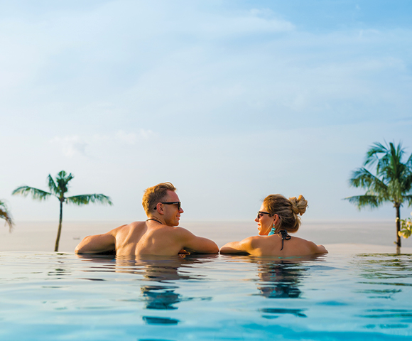 A man and woman relax in an infinity pool, facing each other, with palm trees and a clear sky in the background. The scene is calm and tropical.