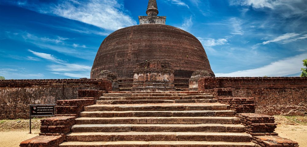Ancient brick stupa with a domed structure and stone steps leading up to it, under a bright blue sky at Polonnaruwa in Sri Lanka.