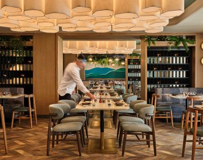 A modern restaurant interior with a waiter setting a table. The space features geometric ceiling lights, wooden accents, and green plants, creating a warm, inviting atmosphere.