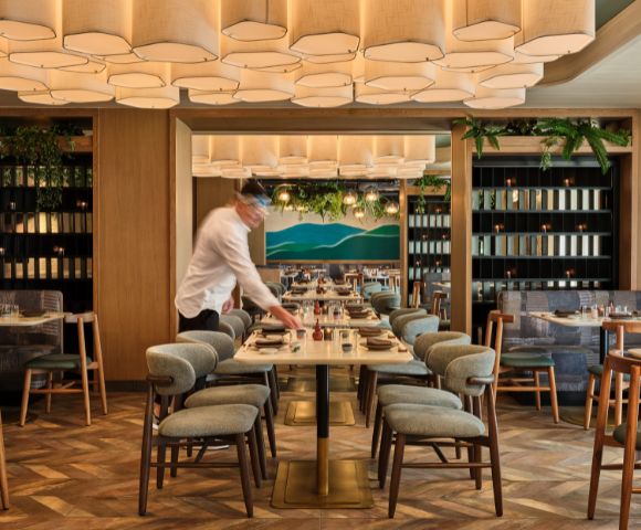 A modern restaurant interior with a waiter setting a table. The space features geometric ceiling lights, wooden accents, and green plants, creating a warm, inviting atmosphere.