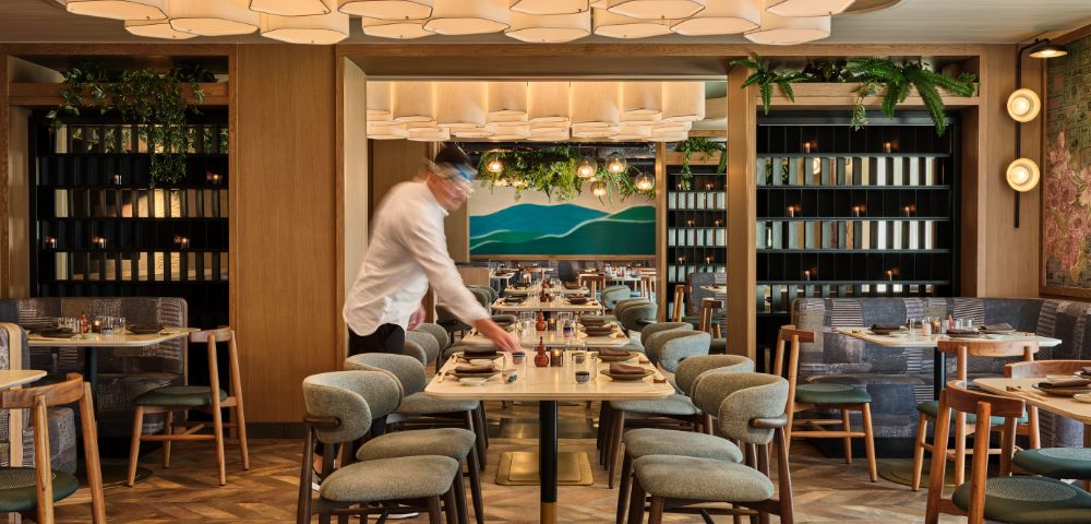 A modern restaurant interior with a waiter setting a table. The space features geometric ceiling lights, wooden accents, and green plants, creating a warm, inviting atmosphere.