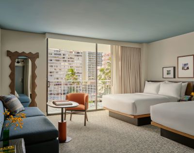 Modern hotel room with two beds, a blue sofa, and a small table with an orange chair. Sliding glass doors open to a balcony with city views and palm trees.