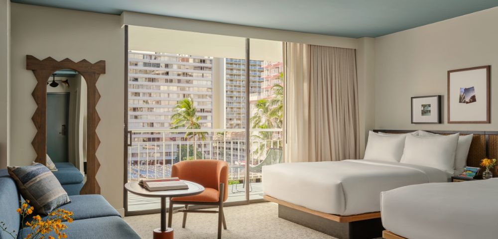 Modern hotel room with two beds, a blue sofa, and a small table with an orange chair. Sliding glass doors open to a balcony with city views and palm trees.