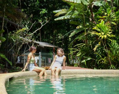 Two people sit at the edge of a serene outdoor pool surrounded by lush greenery. They appear relaxed and are engaged in conversation under soft daylight.