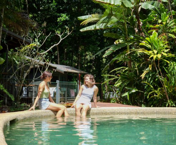 Two people sit at the edge of a serene outdoor pool surrounded by lush greenery. They appear relaxed and are engaged in conversation under soft daylight.