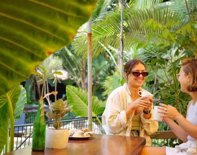 Two women sit at a wooden table in a lush, tropical garden, smiling and holding mugs. Large green leaves frame the relaxed, cheerful scene.