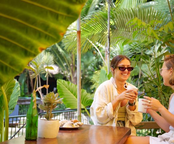 Two women sit at a wooden table in a lush, tropical garden, smiling and holding mugs. Large green leaves frame the relaxed, cheerful scene.