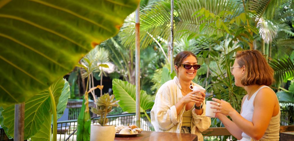 Two women sit at a wooden table in a lush, tropical garden, smiling and holding mugs. Large green leaves frame the relaxed, cheerful scene.