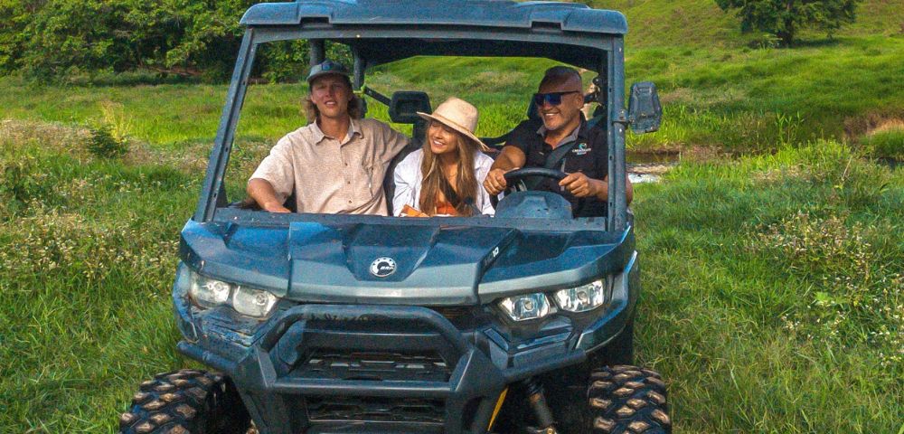 A group of three people enjoys an off-road adventure in a blue ATV, surrounded by lush green hills and clear skies, conveying excitement and joy.