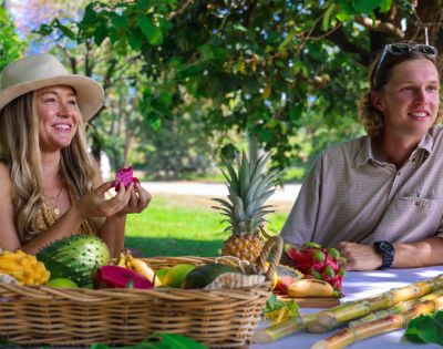 A smiling woman and man enjoy a picnic under a tree with a basket of tropical fruits including pineapple and dragon fruit, conveying a happy, relaxed mood.