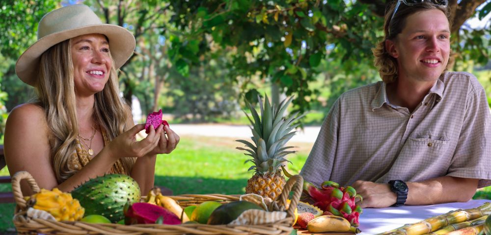 A smiling woman and man enjoy a picnic under a tree with a basket of tropical fruits including pineapple and dragon fruit, conveying a happy, relaxed mood.
