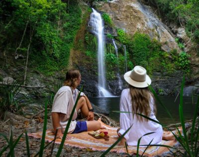 A couple in casual summer attire and a hat sit on a blanket by a waterfall in a lush forest, enjoying a peaceful picnic and a scenic view.