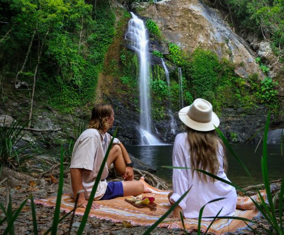 A couple in casual summer attire and a hat sit on a blanket by a waterfall in a lush forest, enjoying a peaceful picnic and a scenic view.