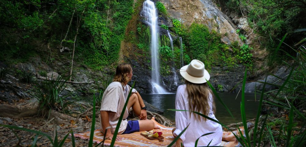 A couple in casual summer attire and a hat sit on a blanket by a waterfall in a lush forest, enjoying a peaceful picnic and a scenic view.
