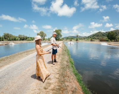 A woman and man fish at a serene pond under a blue sky with scattered clouds. They're on a grassy path, creating a relaxed and peaceful atmosphere.