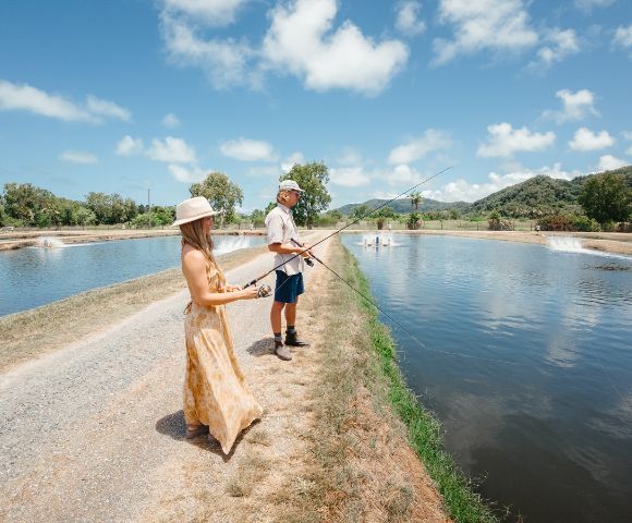 A woman and man fish at a serene pond under a blue sky with scattered clouds. They're on a grassy path, creating a relaxed and peaceful atmosphere.