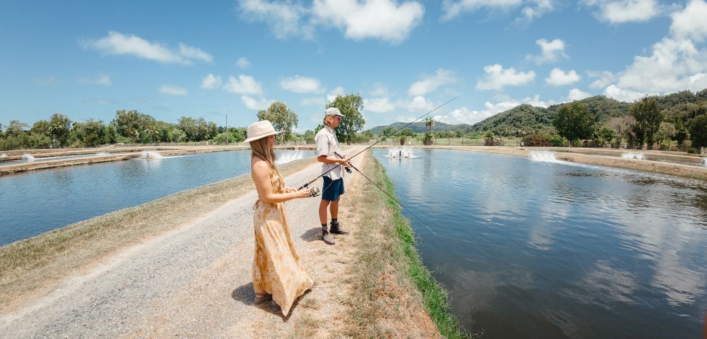 A woman and man fish at a serene pond under a blue sky with scattered clouds. They're on a grassy path, creating a relaxed and peaceful atmosphere.