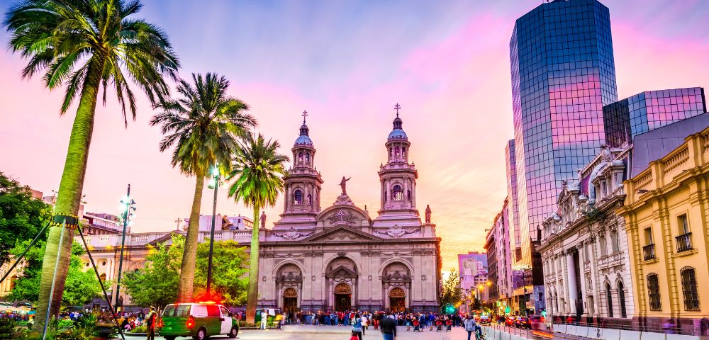 Vibrant cityscape with a historic cathedral framed by tall palm trees at sunset. Warm hues fill the sky, while bustling people create a lively atmosphere.