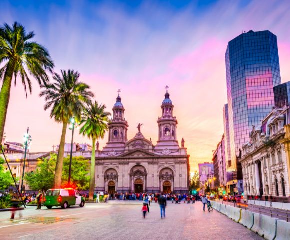 Vibrant cityscape with a historic cathedral framed by tall palm trees at sunset. Warm hues fill the sky, while bustling people create a lively atmosphere.
