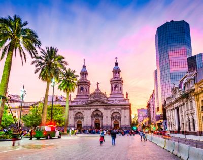 Vibrant cityscape with a historic cathedral framed by tall palm trees at sunset. Warm hues fill the sky, while bustling people create a lively atmosphere.