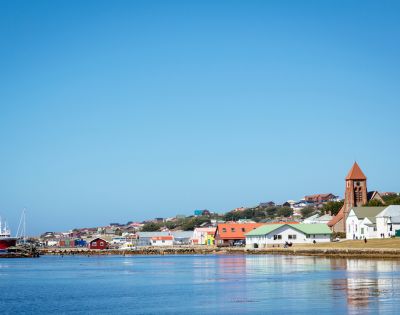 Coastal town with colorful houses and a red-roofed church by the shore. A red fishing boat is docked near the pier under a clear blue sky.