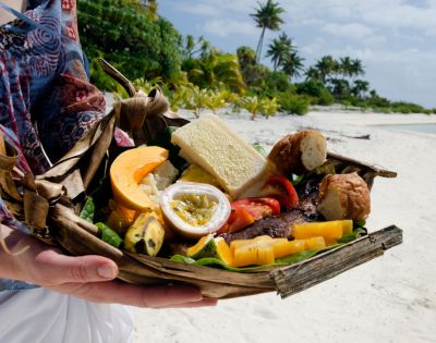 A person holds a woven basket filled with tropical fruits, bread, and grilled meat. The backdrop is a sunny beach with palm trees, conveying a relaxed, vacation vibe.