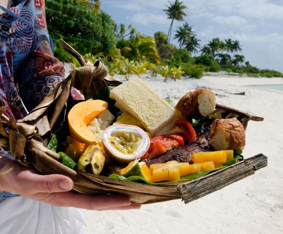 A person holds a woven basket filled with tropical fruits, bread, and grilled meat. The backdrop is a sunny beach with palm trees, conveying a relaxed, vacation vibe.