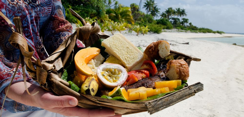 A person holds a woven basket filled with tropical fruits, bread, and grilled meat. The backdrop is a sunny beach with palm trees, conveying a relaxed, vacation vibe.