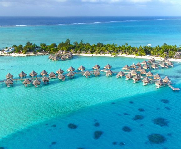 Aerial view of a tropical island with overwater bungalows and lush greenery surrounded by clear turquoise ocean, conveying a serene and idyllic atmosphere.