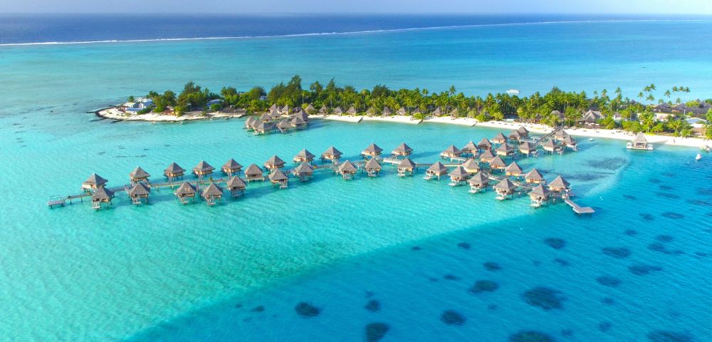 Aerial view of a tropical island with overwater bungalows and lush greenery surrounded by clear turquoise ocean, conveying a serene and idyllic atmosphere.