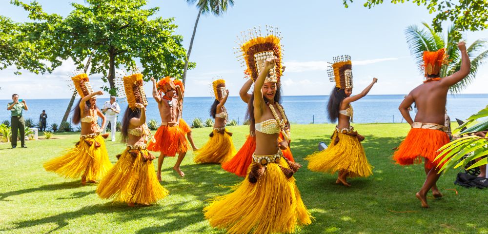 A group of dancers in vibrant, traditional Polynesian attire perform energetically on lush grass, with palm trees and ocean in the sunny background.